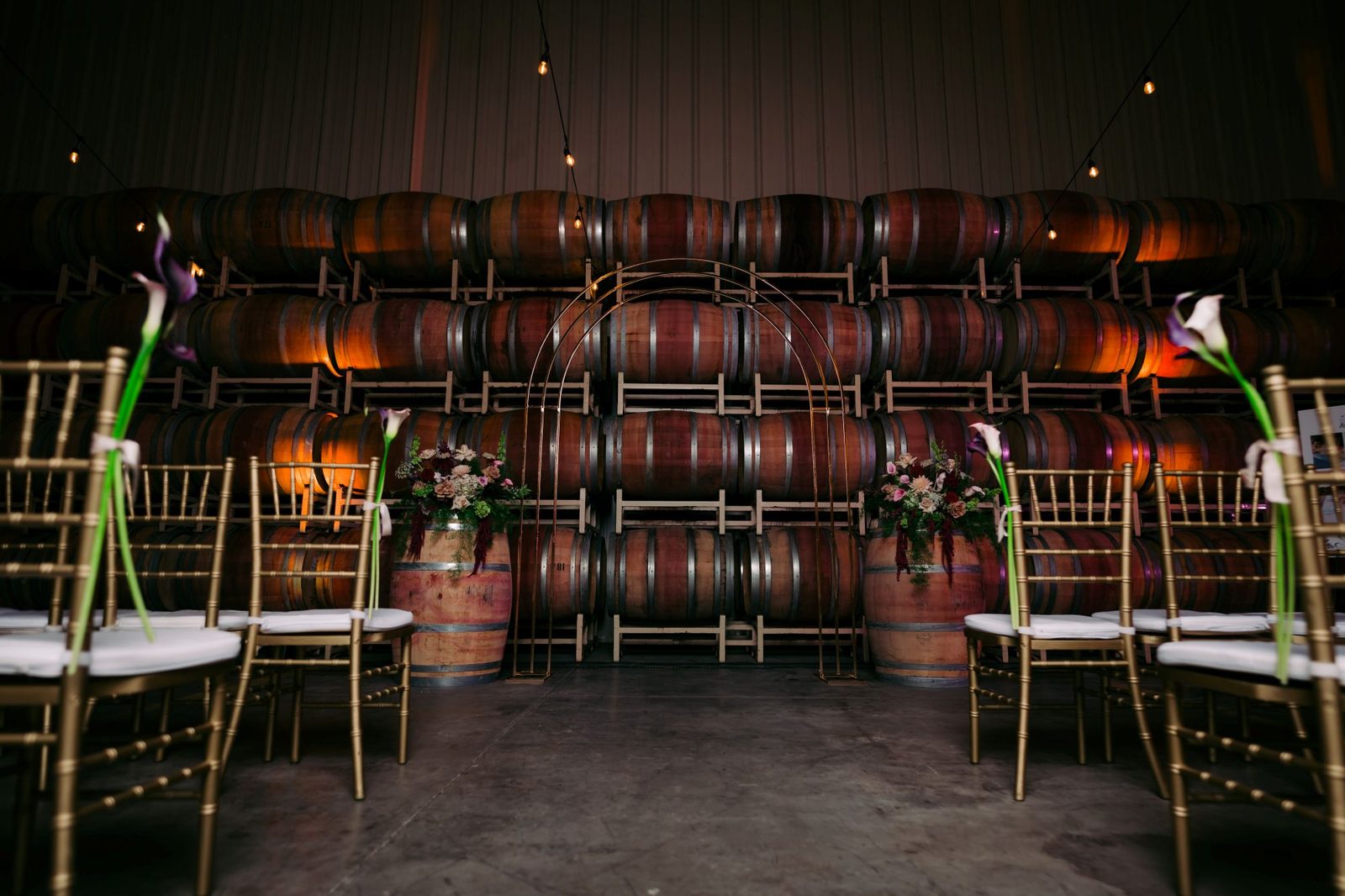 Wedding ceremony set up in The Barrel Hall with gold chiavari chairs, floral arrangements, and a gold arch against a wall of wine barrels