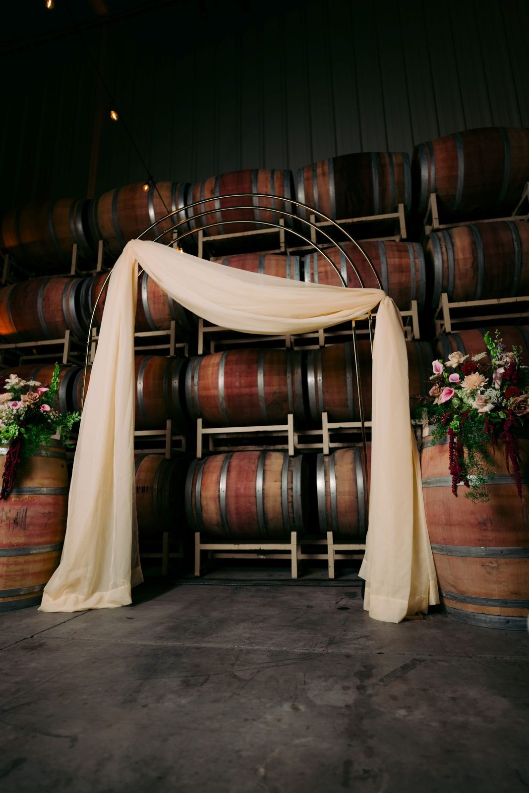 Draped ceremony arch with ivory fabric and floral accents in front of stacked wine barrels at The Barrel Hall