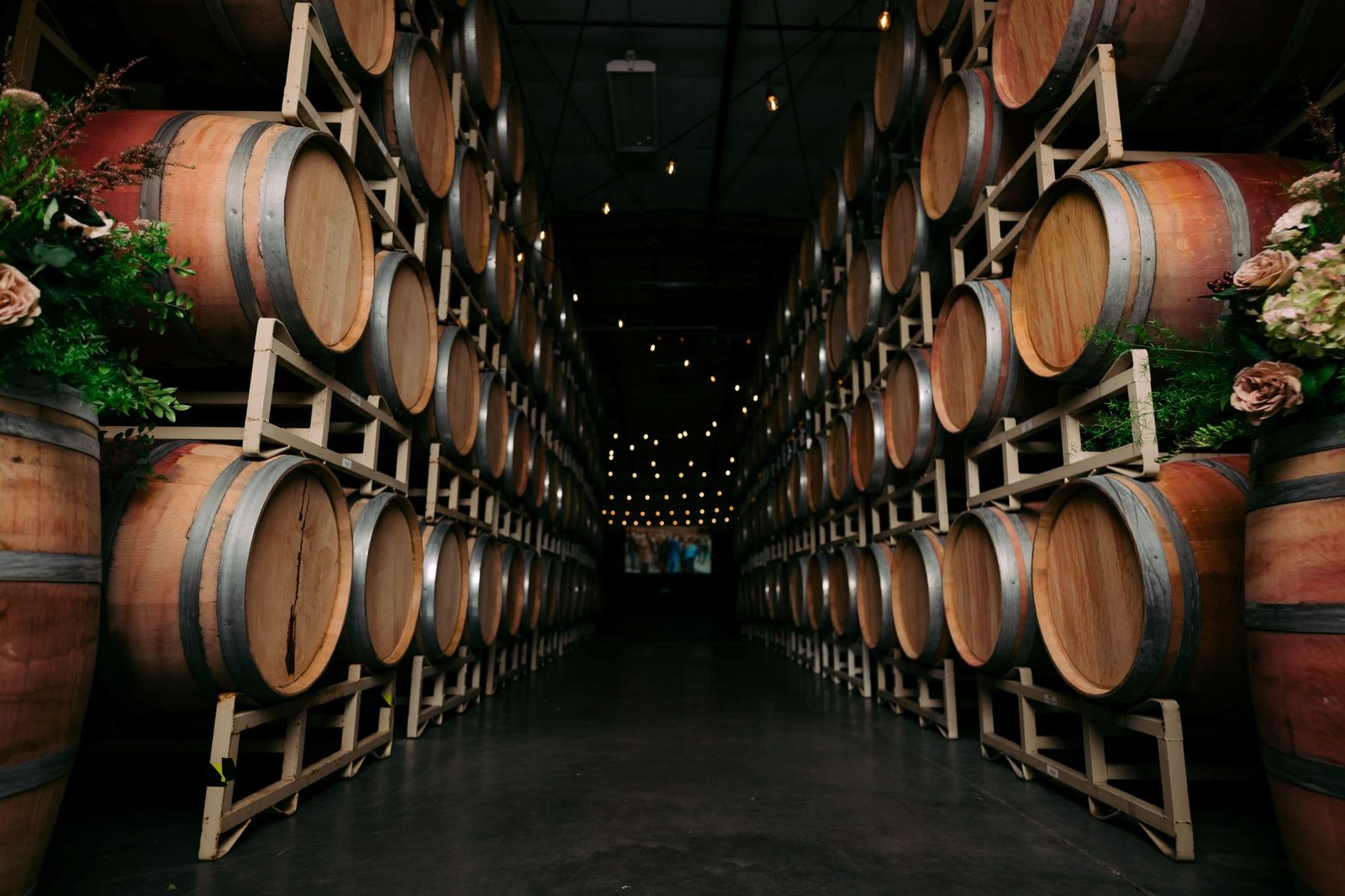 Aisle between towering rows of wine barrels lit by string lights at The Barrel Hall at Goose Ridge Estate