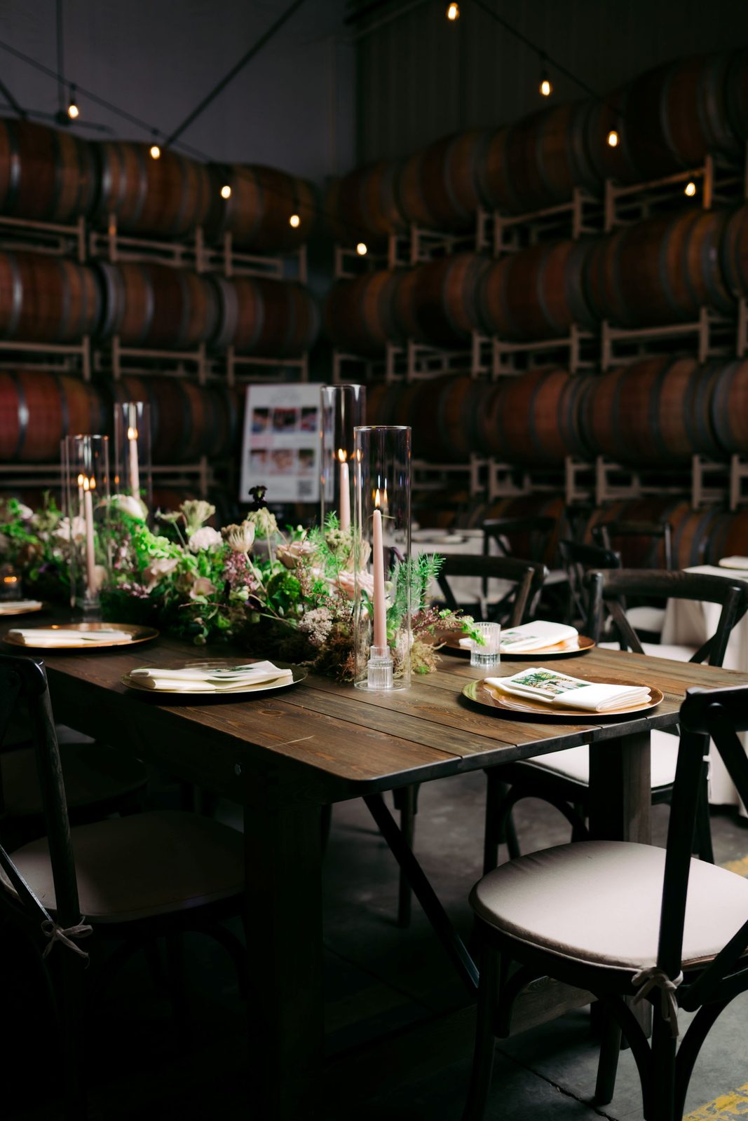 Farmhouse tables set for a reception with candles and greenery among the wine barrels at The Barrel Hall