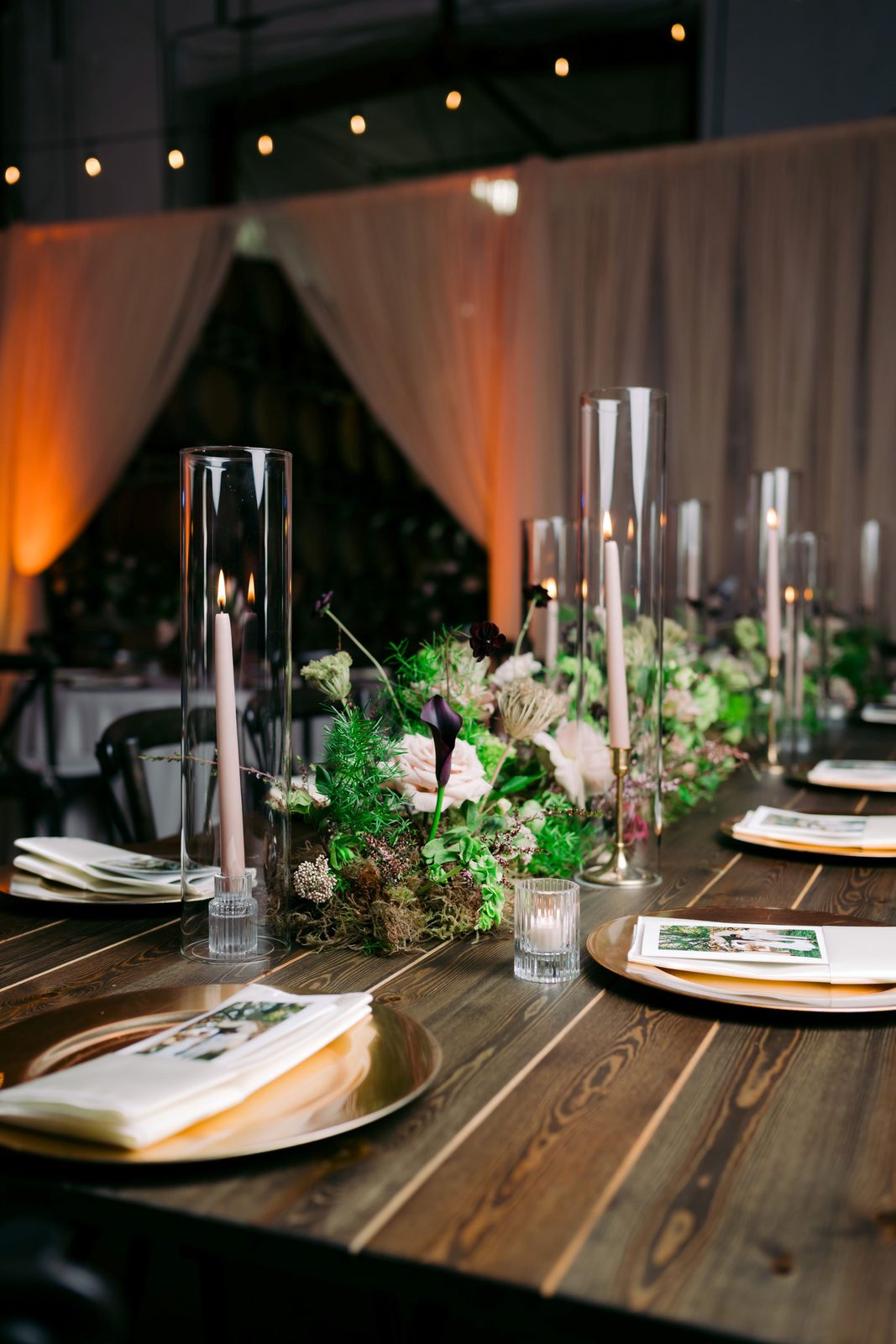 Reception table detail with blush taper candles, gold chargers, and greenery at The Barrel Hall