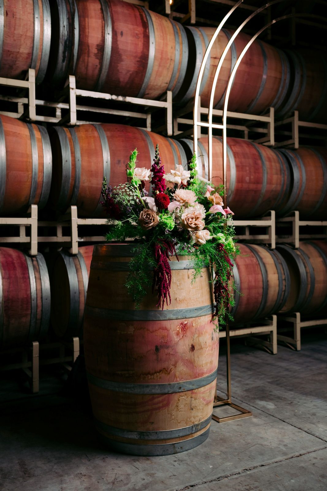 Lush floral arrangement atop a wine barrel beside a gold arch at The Barrel Hall